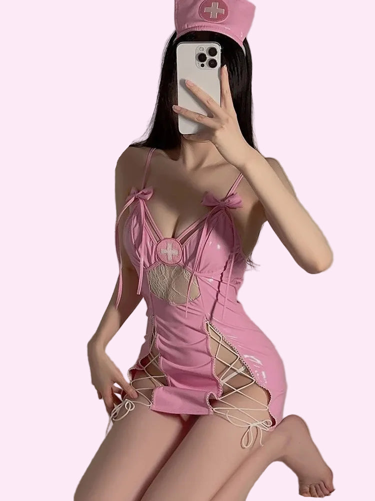 A woman takes a mirror selfie wearing a pink nurse-themed mini dress with lace-up details and a matching pink nurse hat. The background is a plain indoor wall and bed.