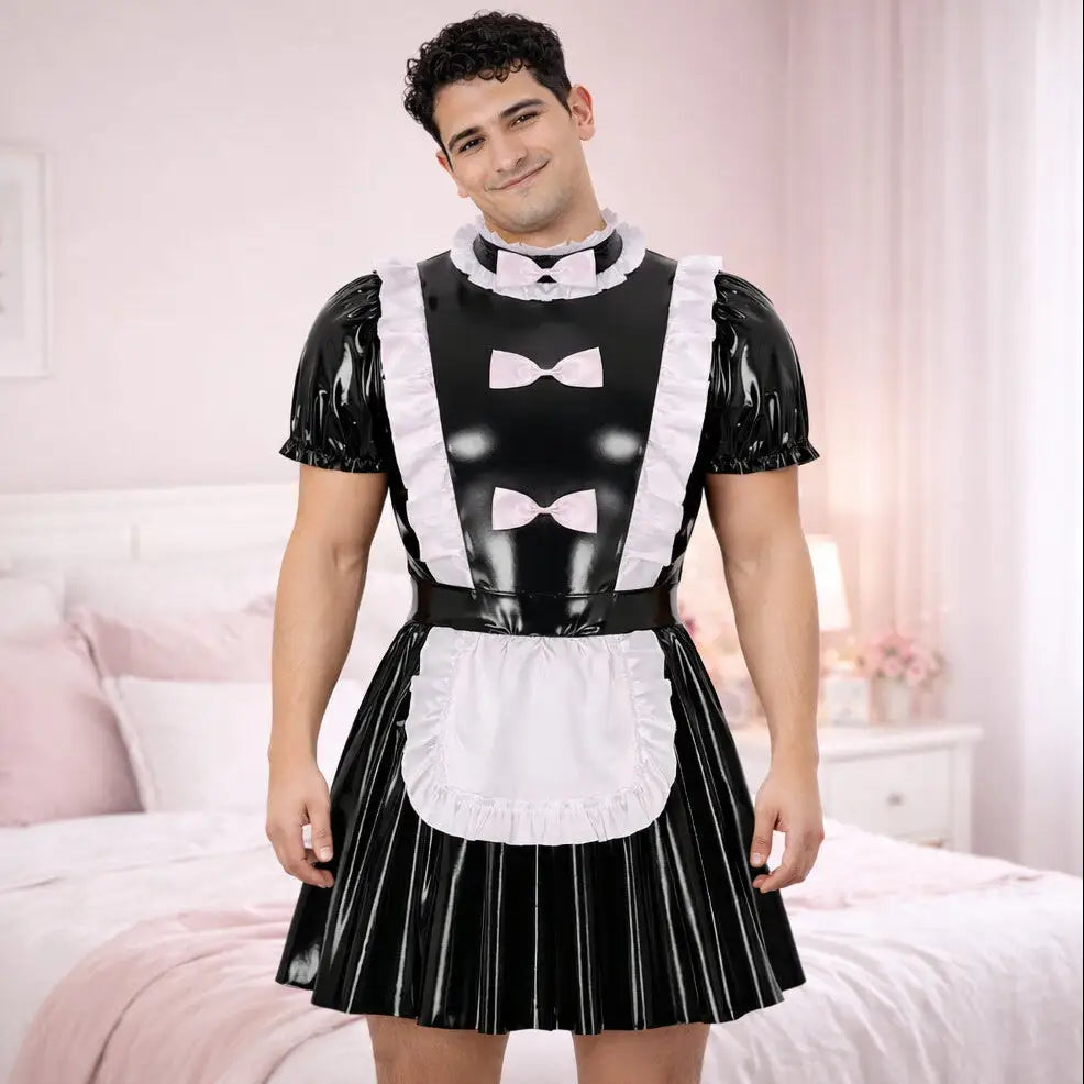 Man wearing a black latex dress with white apron and bow ties in a bedroom setting.
